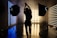 A silhouetted person stands in a photography studio, framed by two large studio lights on stands. The room is dimly lit with linear blue and yellow lights creating a contrasting background. Smoke or fog adds an atmospheric effect, with the floor reflecting the lights.