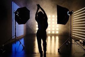A silhouetted person stands in a photography studio, framed by two large studio lights on stands. The room is dimly lit with linear blue and yellow lights creating a contrasting background. Smoke or fog adds an atmospheric effect, with the floor reflecting the lights.