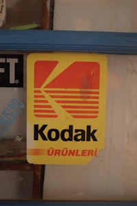 A vintage Kodak sign with a yellow background and red text is displayed behind a glass window. The sign has a large stylized 'K' logo and the word 'Kodak' in black letters, followed by 'ÜRÜNLERİ' in red at the bottom. The edges show signs of wear, indicating age.