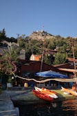 A tranquil coastal scene features kayaks resting by the water in the foreground. A rustic wooden building is partially covered by vibrant bougainvillea. Above, a hill is topped with an ancient stone fortress displaying a prominent red flag, surrounded by lush greenery under a clear blue sky.