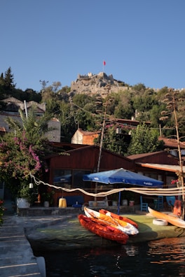 A tranquil coastal scene features kayaks resting by the water in the foreground. A rustic wooden building is partially covered by vibrant bougainvillea. Above, a hill is topped with an ancient stone fortress displaying a prominent red flag, surrounded by lush greenery under a clear blue sky.