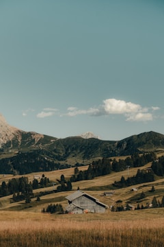 A rustic wooden barn is nestled amidst rolling hills covered with lush grass and scattered trees. In the background, there are tall, rugged mountains under a clear, blue sky with a few fluffy clouds.