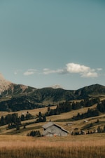 A rustic wooden barn is nestled amidst rolling hills covered with lush grass and scattered trees. In the background, there are tall, rugged mountains under a clear, blue sky with a few fluffy clouds.