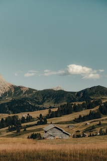 A rustic wooden barn is nestled amidst rolling hills covered with lush grass and scattered trees. In the background, there are tall, rugged mountains under a clear, blue sky with a few fluffy clouds.