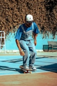 A person wearing a blue shirt and overalls is skateboarding on a court with blue and brown colors. The individual is wearing a white baseball cap with a black logo and is balancing on the skateboard. In the background, there's a bench and a blue fence with a dense tree or bush casting shadows.
