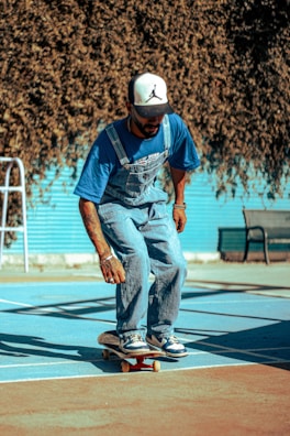 A person wearing a blue shirt and overalls is skateboarding on a court with blue and brown colors. The individual is wearing a white baseball cap with a black logo and is balancing on the skateboard. In the background, there's a bench and a blue fence with a dense tree or bush casting shadows.