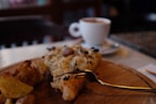 A wooden board holds a slice of pizza topped with cheese and herbs, accompanied by potato wedges. In the background, a white cup of coffee sits on a saucer, slightly blurred.