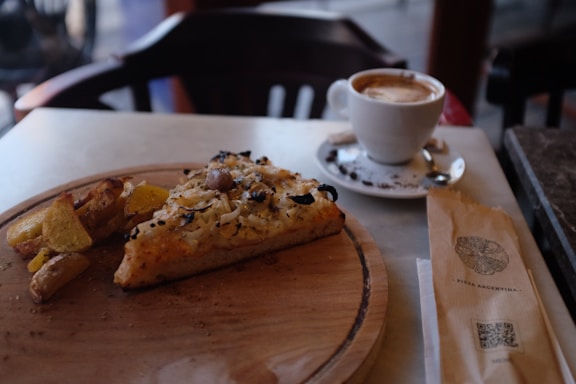 A slice of pizza topped with cheese, olives, and mushrooms is placed on a wooden platter. Beside it, there are crispy roasted potato wedges. In the background, a cup of coffee with a saucer and spoon on a white table. A paper bag with the inscription 'Pizza Argentina' and a QR code is visible next to the plate.