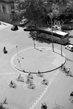 A group of cyclists navigate a roundabout in a city street, surrounded by cars, a bus, and pedestrians near storefronts. There are trees casting shadows and the scene is captured in black and white.