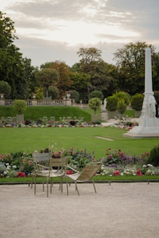A tranquil garden setting features a pair of empty chairs and a lounge chair on a gravel path. The lush green lawn is adorned with colorful flowerbeds, including reds, pinks, whites, and purples. An obelisk stands in the background against a backdrop of mature trees and a partly cloudy sky, hinting at dusk.