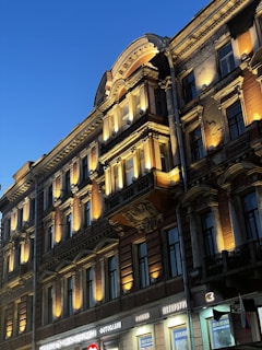 An ornate, multi-story building showcasing detailed architectural elements such as columns and decorative moldings. Warm yellow lighting highlights the intricate designs on the façade. A vibrant blue sky provides a contrasting backdrop, while signs with Cyrillic text are visible at the ground level.