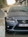 A close-up view of a gray Lexus car undergoing a wash, with water spraying from the left side. The surface of the car is wet with droplets, and the background shows a blurred setting with buildings and greenery.