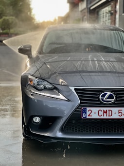 A close-up view of a gray Lexus car undergoing a wash, with water spraying from the left side. The surface of the car is wet with droplets, and the background shows a blurred setting with buildings and greenery.