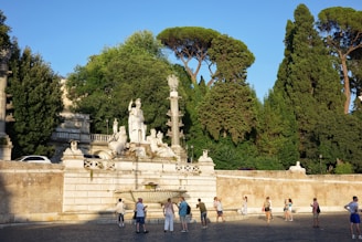 A historical monument with classical statues is set against a backdrop of tall, lush trees. People are gathered around, some standing, others appearing to be tourists, observing the monument.
