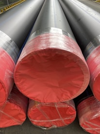 Large cylindrical pipes with black bodies and red ends are stacked and wrapped in plastic. They are positioned on a paved surface near a fence with yellow markers.