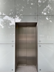 An elevator with metallic doors is set into a modern exterior with frosted glass panels. The reflection of surrounding trees and a building is faintly visible on the glass, giving a somewhat ethereal appearance.