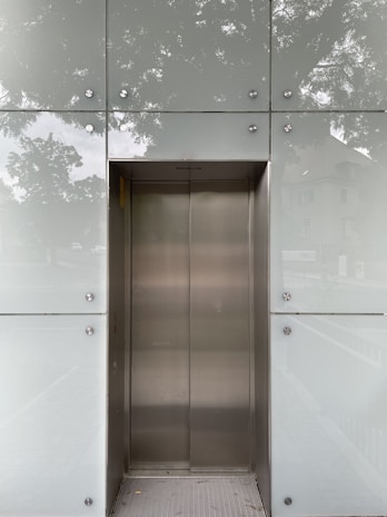 An elevator with metallic doors is set into a modern exterior with frosted glass panels. The reflection of surrounding trees and a building is faintly visible on the glass, giving a somewhat ethereal appearance.