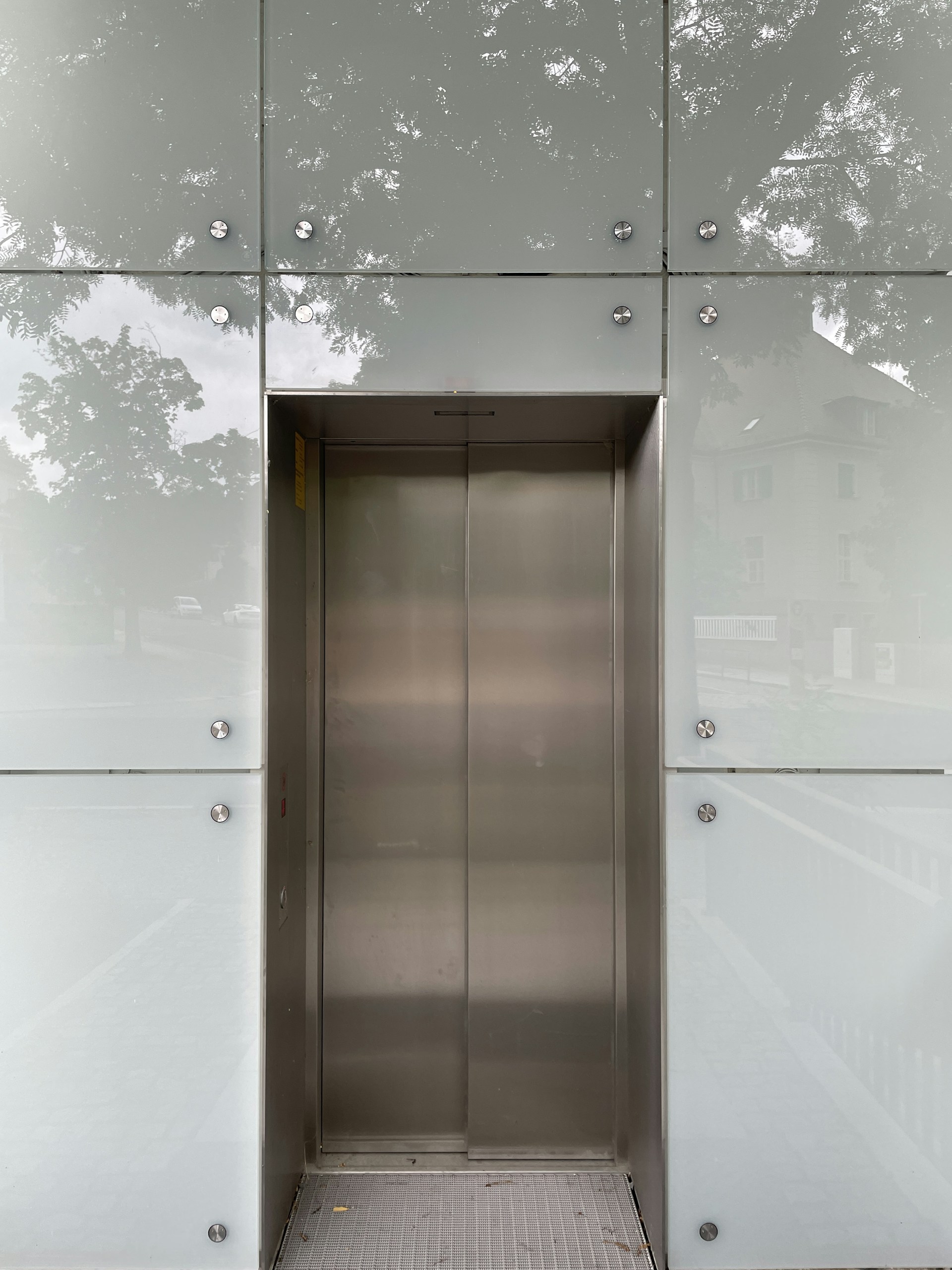 An elevator with metallic doors is set into a modern exterior with frosted glass panels. The reflection of surrounding trees and a building is faintly visible on the glass, giving a somewhat ethereal appearance.