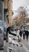 A street scene featuring people browsing artwork displayed on steps. The setting includes columns covered in graffiti and trees in the background. Several pedestrians, including a child and their guardian, walk by, and an artist arranges paintings on the sidewalk.