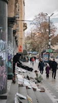 A street scene featuring people browsing artwork displayed on steps. The setting includes columns covered in graffiti and trees in the background. Several pedestrians, including a child and their guardian, walk by, and an artist arranges paintings on the sidewalk.