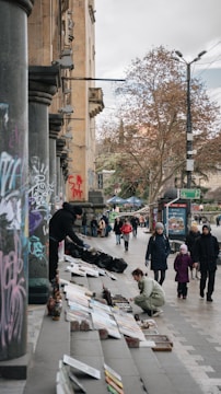 A street scene featuring people browsing artwork displayed on steps. The setting includes columns covered in graffiti and trees in the background. Several pedestrians, including a child and their guardian, walk by, and an artist arranges paintings on the sidewalk.