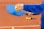 A person wearing a blue sweatshirt holds a blue pickleball paddle and a yellow pickleball. The setting is an outdoor court with a few additional yellow pickleballs lying on the ground. The court surface appears to be made of clay.