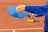 A person wearing a blue sweatshirt holds a blue pickleball paddle and a yellow pickleball. The setting is an outdoor court with a few additional yellow pickleballs lying on the ground. The court surface appears to be made of clay.