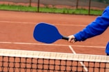 A person wearing a blue long-sleeve shirt holds a blue pickleball paddle over a net on a clay court.