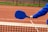 A person wearing a blue long-sleeve shirt holds a blue pickleball paddle over a net on a clay court.