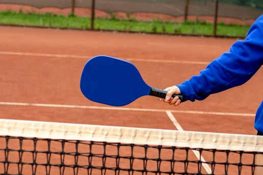 A person wearing a blue long-sleeve shirt holds a blue pickleball paddle over a net on a clay court.