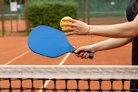 A person holding a blue paddle and a yellow pickleball over a tennis court's net. The court surface is orange clay, and there is greenery in the background.