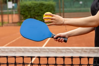 A friendly tennis ball resting on a pickleball paddle with a blurred court background.