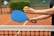 A person holding a blue paddle and a yellow pickleball over a tennis court's net. The court surface is orange clay, and there is greenery in the background.