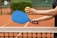 A person holding a blue paddle and a yellow pickleball over a tennis court's net. The court surface is orange clay, and there is greenery in the background.