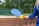 A person is holding a blue paddle and a yellow perforated ball next to a net on what appears to be a sports court. The background features a fence and greenery, suggesting an outdoor setting. The person is wearing a blue long-sleeved top.