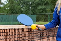 A person is holding a blue paddle and a yellow perforated ball next to a net on what appears to be a sports court. The background features a fence and greenery, suggesting an outdoor setting. The person is wearing a blue long-sleeved top.
