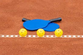 Two blue paddles and three yellow perforated balls are placed on a brown clay court, with a white dashed line running horizontally across the image.