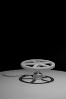 A large industrial valve with a wheel-shaped handle is attached to a flat surface. The valve and handle are prominently featured against a black background, casting distinct shadows on the surface. The simplicity and stark contrast give a dramatic effect to the mechanical object.