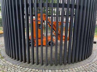 An orange construction lift is enclosed within a circular structure made of vertical black metal bars. The bars form a cage-like pattern around the lift. The ground is paved with cobblestones, and there is greenery visible in the background.