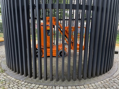 An orange construction lift is enclosed within a circular structure made of vertical black metal bars. The bars form a cage-like pattern around the lift. The ground is paved with cobblestones, and there is greenery visible in the background.