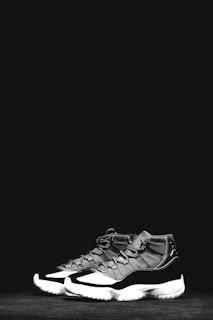 A pair of stylish, black and white athletic sneakers with a high-top design are placed against a solid black background. The shoes feature a glossy finish and intricate lacing details.