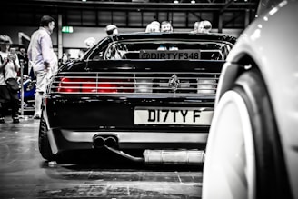 A sleek black sports car is displayed at an indoor automotive event with several people around it, some seemingly taking photos. The car's rear end, including the license plate and distinctive tail lights, is prominently featured. The reflections and lighting add a polished, high-end feel to the scene.