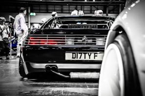 A sleek black sports car is displayed at an indoor automotive event with several people around it, some seemingly taking photos. The car's rear end, including the license plate and distinctive tail lights, is prominently featured. The reflections and lighting add a polished, high-end feel to the scene.