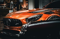 A sleek, orange sports car prominently displayed with its front view captured, showcasing its aggressive grille and shiny headlight. The car is indoors with a blurred background, possibly in a showroom or mall, with people walking by.