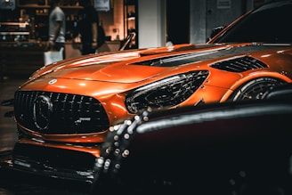 A sleek, orange sports car prominently displayed with its front view captured, showcasing its aggressive grille and shiny headlight. The car is indoors with a blurred background, possibly in a showroom or mall, with people walking by.