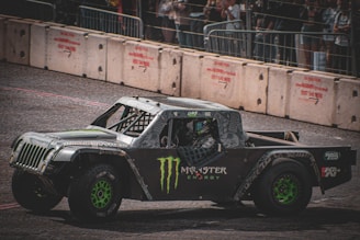 A rugged off-road racing truck with a bold design featuring logos and green wheels is driving on a track. The vehicle has a reinforced exterior and is surrounded by concrete barriers. Spectators are visible behind a fence, watching the event.