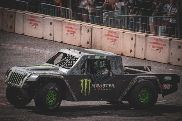 A rugged off-road racing truck with a bold design featuring logos and green wheels is driving on a track. The vehicle has a reinforced exterior and is surrounded by concrete barriers. Spectators are visible behind a fence, watching the event.