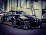 A sleek black sports car with a shiny finish is displayed indoors, featuring decals and a prominent front bumper. The car is positioned on a black floor with industrial-style architecture in the background, and banners can be seen partially in the vicinity.