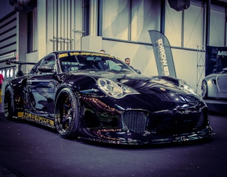 A sleek black sports car with a shiny finish is displayed indoors, featuring decals and a prominent front bumper. The car is positioned on a black floor with industrial-style architecture in the background, and banners can be seen partially in the vicinity.