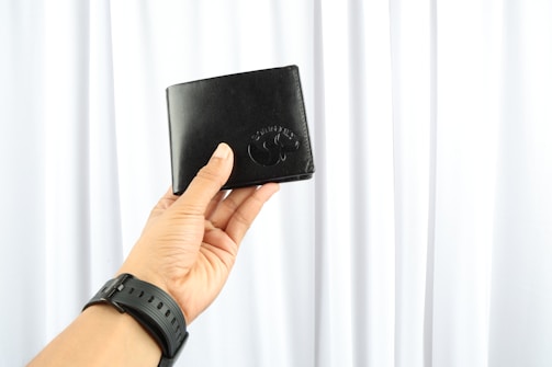 A hand is holding a black wallet against a background of white curtains. The wallet is square-shaped and appears to be made of leather. The person is wearing a black wristwatch.